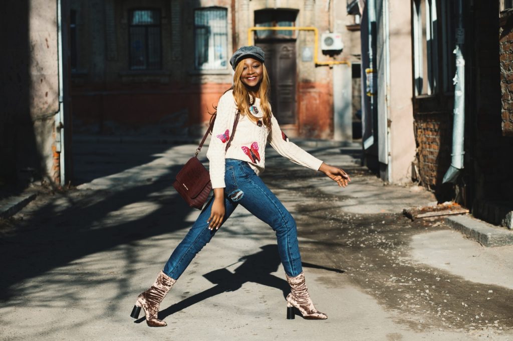 femme marchant dans la rue portant un pull blanc avec des papillons rouges un jean et des bottines en velours dorée un sac sur l'épaule et une casquette grise